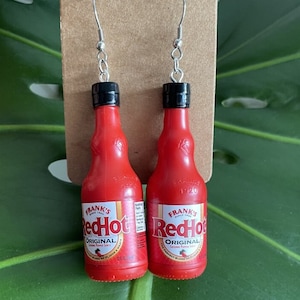 Op de afbeelding: Een paar rode hete sausfles oorringen. De oorringen zijn gemaakt van rood plastic en hebben een zwarte dop. De fles heeft de tekst "Frank's RedHot Original Cayenne Pepper Sauce" op het etiket.