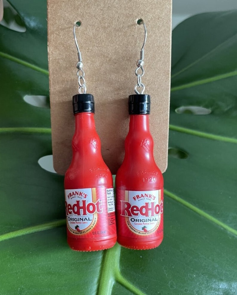 Op de afbeelding: Een paar rode hete sausfles oorringen. De oorringen zijn gemaakt van rood plastic en hebben een zwarte dop. De fles heeft de tekst "Frank's RedHot Original Cayenne Pepper Sauce" op het etiket.