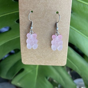 K&ouml;nnte beinhalten: Ein Paar Ohrringe mit pinken Gummib&auml;rchen-Anh&auml;ngern. Die Ohrringe sind aus silbernem Metall und haben einen Hakenverschluss.