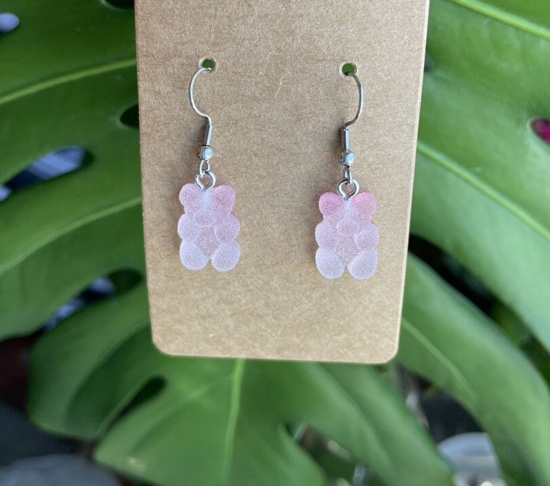 K&ouml;nnte beinhalten: Ein Paar Ohrringe mit pinken Gummib&auml;rchen-Anh&auml;ngern. Die Ohrringe sind aus silbernem Metall und haben einen Hakenverschluss.