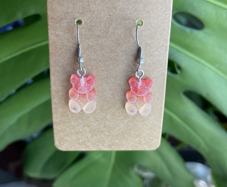 K&ouml;nnte beinhalten: Ein Paar silberne Ohrh&auml;nger mit einem Gummib&auml;rchen-Anh&auml;nger. Das Gummib&auml;rchen ist ein Farbverlauf von Rot, Orange und Gelb.