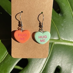 May include: A pair of heart-shaped earrings with silver hooks. The hearts are made of red and green plastic and have the words "Hug Me" written on them in pink.