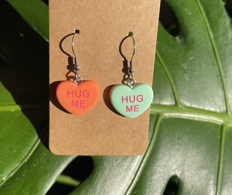 May include: A pair of heart-shaped earrings with silver hooks. The hearts are made of red and green plastic and have the words "Hug Me" written on them in pink.