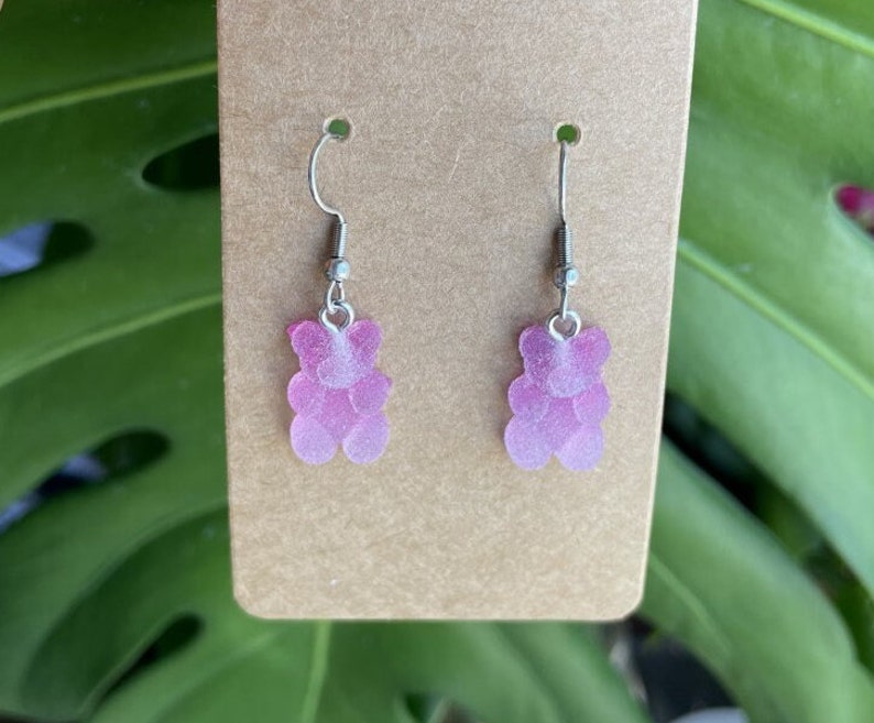 K&ouml;nnte beinhalten: Ein Paar Ohrringe mit einem rosa Gummib&auml;rchen-Anh&auml;nger. Die Ohrringe haben silberne Haken.