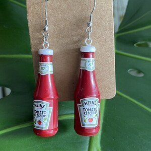 Op de afbeelding: Een paar rode ketchupfles-oorbellen met zilveren haken. De flessen hebben een wit etiket met de tekst "HEINZ TOMATO KETCHUP 57 VARIETIES" en een rode tomaat.