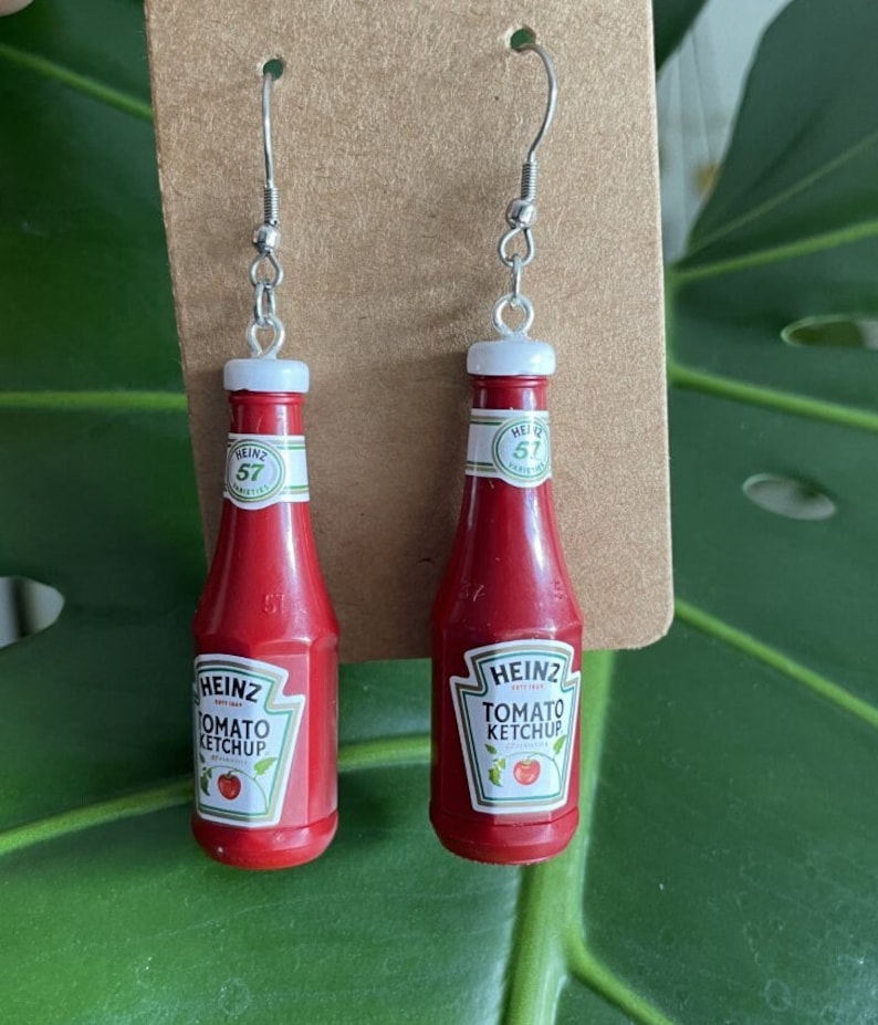 Op de afbeelding: Een paar rode ketchupfles-oorbellen met zilveren haken. De flessen hebben een wit etiket met de tekst "HEINZ TOMATO KETCHUP 57 VARIETIES" en een rode tomaat.