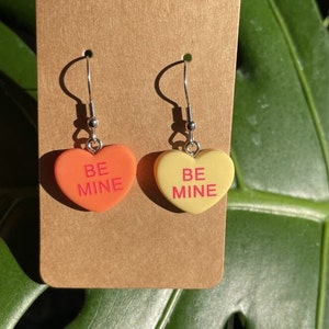 May include: A pair of heart-shaped earrings with silver hooks. The hearts are made of plastic and have the words "Be Mine" written on them. One heart is orange and the other is yellow.