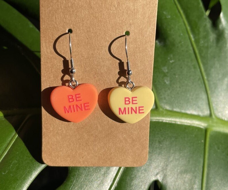 May include: A pair of heart-shaped earrings with silver hooks. The hearts are made of plastic and have the words "Be Mine" written on them. One heart is orange and the other is yellow.