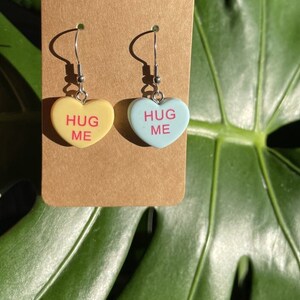 May include: A pair of silver dangle earrings with heart-shaped charms. The charms are in the shape of candy hearts and say "Hug Me" in pink lettering. One charm is yellow and the other is light blue.