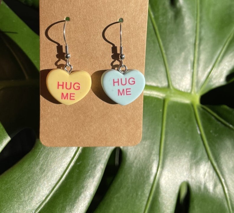 May include: A pair of silver dangle earrings with heart-shaped charms. The charms are in the shape of candy hearts and say "Hug Me" in pink lettering. One charm is yellow and the other is light blue.