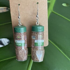 Op de afbeelding: Een paar oorbellen in de vorm van kleine kruidenpotjes met groene deksels. De potjes zijn gelabeld met "McCormick Organic Thyme".