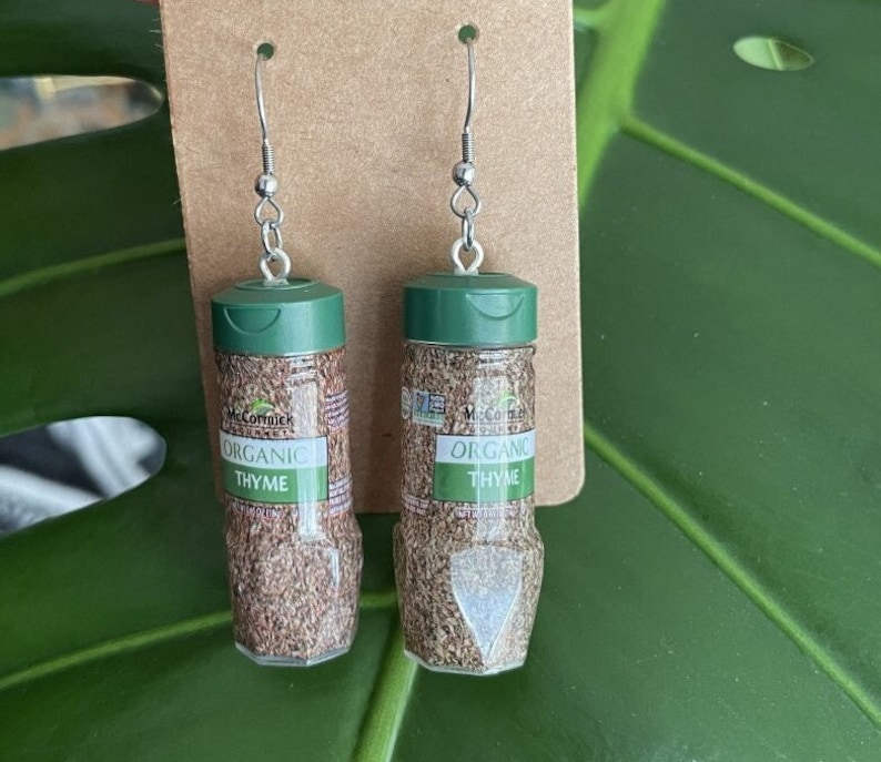 Op de afbeelding: Een paar oorbellen in de vorm van kleine kruidenpotjes met groene deksels. De potjes zijn gelabeld met "McCormick Organic Thyme".