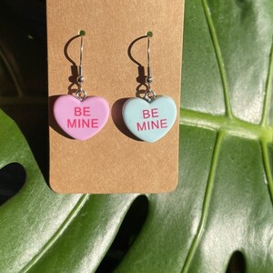 May include: A pair of dangle earrings featuring heart-shaped charms with the text "BE MINE" in pink lettering. One charm is pink and the other is light blue.