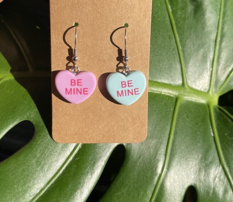 May include: A pair of dangle earrings featuring heart-shaped charms with the text "BE MINE" in pink lettering. One charm is pink and the other is light blue.
