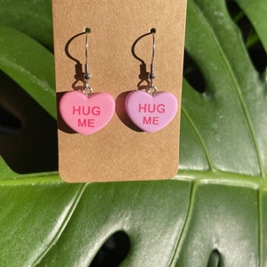 May include: A pair of pink heart-shaped earrings with the text "HUG ME" on each heart. The earrings are on a brown card.