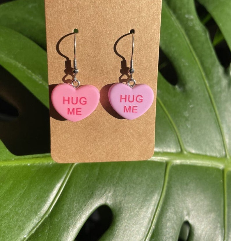 May include: A pair of pink heart-shaped earrings with the text "HUG ME" on each heart. The earrings are on a brown card.