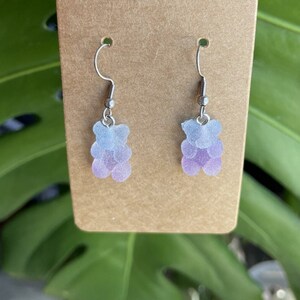 K&ouml;nnte beinhalten: Ein Paar silberne Ohrh&auml;nger mit einem hellblauen und violetten Ombre-Gummib&auml;rchen-Anh&auml;nger.