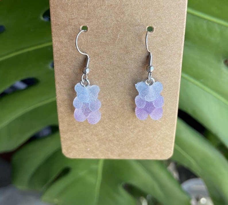 K&ouml;nnte beinhalten: Ein Paar silberne Ohrh&auml;nger mit einem hellblauen und violetten Ombre-Gummib&auml;rchen-Anh&auml;nger.