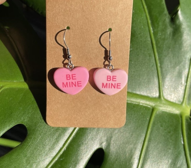 May include: A pair of pink heart-shaped earrings with silver hooks. The hearts have the words "BE MINE" written on them.