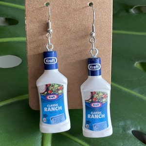 Op de afbeelding: Een paar zilveren oorhangers met miniatuur plastic flesjes Kraft Classic Ranch dressing. De flesjes zijn wit met blauwe en rode accenten en de tekst "Kraft Classic Ranch Dressing" staat op de voorkant.