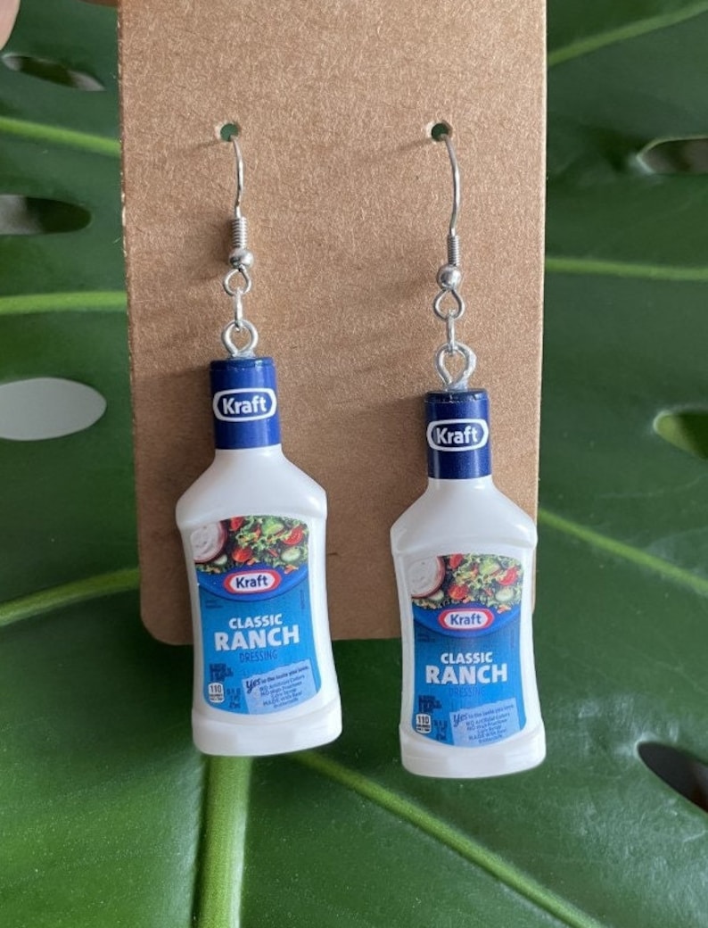 Op de afbeelding: Een paar zilveren oorhangers met miniatuur plastic flesjes Kraft Classic Ranch dressing. De flesjes zijn wit met blauwe en rode accenten en de tekst "Kraft Classic Ranch Dressing" staat op de voorkant.