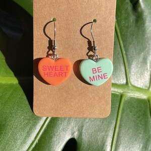 May include: A pair of heart-shaped earrings with silver hooks. The left earring is orange and says "SWEET HEART" in pink lettering. The right earring is green and says "BE MINE" in pink lettering.