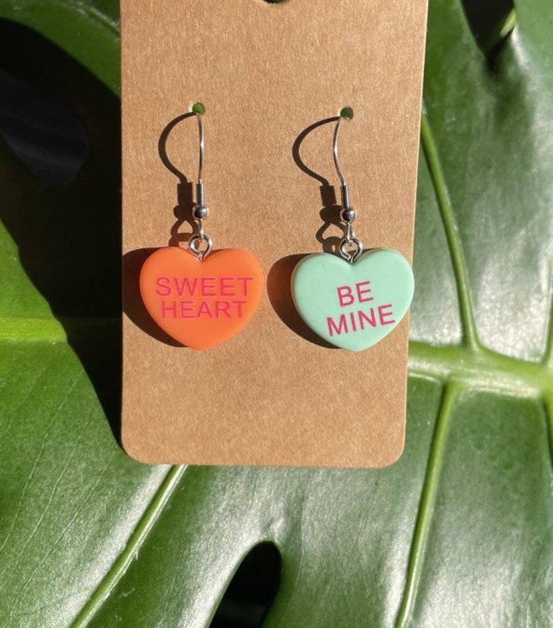 May include: A pair of heart-shaped earrings with silver hooks. The left earring is orange and says "SWEET HEART" in pink lettering. The right earring is green and says "BE MINE" in pink lettering.