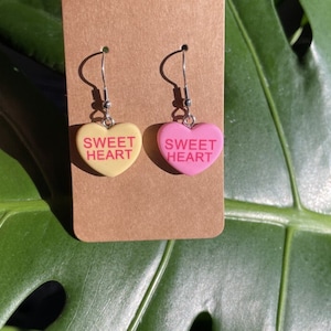 May include: A pair of heart-shaped earrings with the text "SWEET HEART" on each. One earring is yellow and the other is pink.