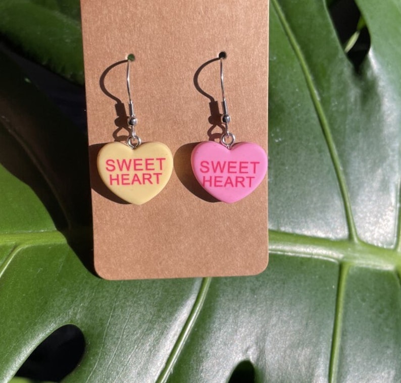 May include: A pair of heart-shaped earrings with the text "SWEET HEART" on each. One earring is yellow and the other is pink.