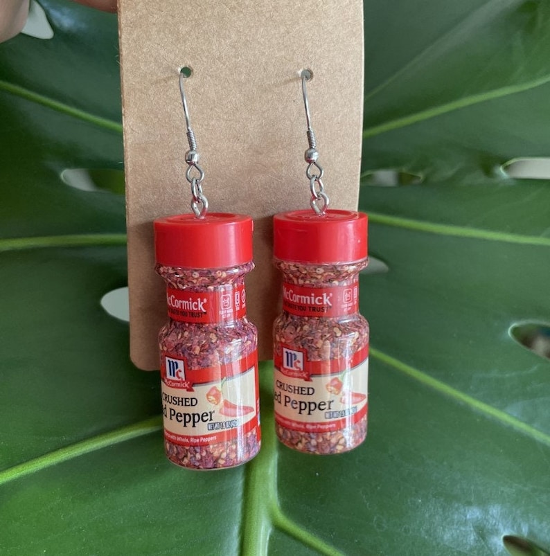 Op de afbeelding: Een paar rode en witte gebroken rode peper oorbellen. De oorbellen zijn gevormd als kleine kruidenpotjes met het McCormick-merklabel en de woorden "Crushed Red Pepper".