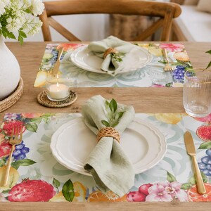 Puede incluir: Una mesa para dos con manteles individuales florales con diseños de frutas y flores. Platos blancos, servilletas verdes y cubiertos dorados están colocados sobre los manteles individuales. Un jarrón con flores blancas y una vela encendida completan el ambiente.