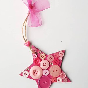 May include: Pink wooden star ornament decorated with pink, red and white buttons. The star has a pink ribbon attached to the top.