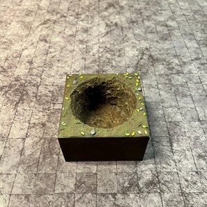 May include: A miniature, square-shaped diorama with a dark brown base and a detailed, textured interior. The top surface is painted green with small, scattered yellow and silver accents, resembling leaves and gems. The interior features a deep, carved-out area.