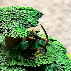 May include: Miniature archer figure in a green tunic and cape, drawing a bow. The figure is positioned on a textured green base, surrounded by a detailed, leafy green tree. The background is a neutral gray.