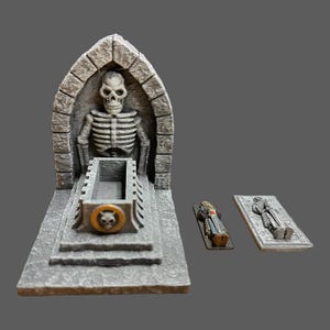May include: A 3D printed miniature tomb with a skeletal figure inside. The tomb has a stone archway and a coffin with a skull emblem. There are two additional miniature coffins on the side.