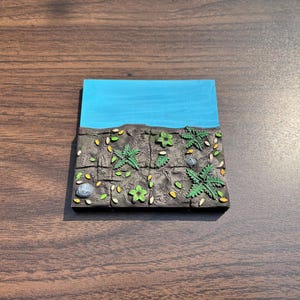 May include: A miniature diorama featuring a landscape with a blue sky, a dark brown earth section, and green plants. The earth section has a textured appearance with small rocks and scattered leaves. The diorama is set on a wooden surface.