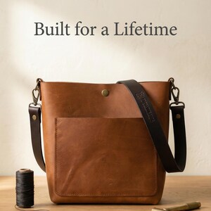 May include: A brown leather tote bag with a front pocket and a black shoulder strap. The bag has a button closure and metal hardware. The text "Built for a Lifetime" is above the bag. A spool of black thread and a small tool are on the table.