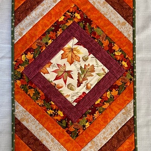 May include: A quilted table runner with a chevron pattern in shades of orange, brown, and white. The center features a burgundy diamond with a cream fabric inset featuring a fall leaf print and the words "gather, give thanks, bless our family".