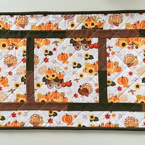 May include: Rectangular quilted table runner with a white background featuring a repeating pattern of pumpkins, sunflowers, and turkeys. The runner has a brown and green border and a central design of brown and green squares.