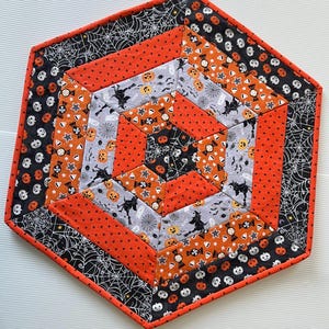 May include: Hexagon-shaped Halloween-themed quilted table topper. The topper features orange, black, and gray fabrics with Halloween patterns like pumpkins, bats, and spiderwebs. The edges are finished with a red binding.