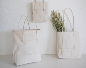 Puede incluir: Tres bolsas de lona de color blanco roto con un diseño sencillo. Las bolsas tienen un solo bolsillo en la parte delantera y están hechas de una lona gruesa y resistente.