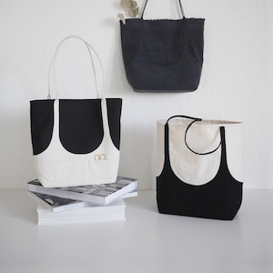 May include: Three tote bags made of black and white canvas. The bags have a unique design with a curved, contrasting color panel on the front. Two of the bags have a small pocket with two gold rings on the front.