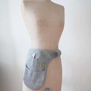 Puede incluir: Un cintur&oacute;n de herramientas de tela gris con un bolsillo y un par de tijeras. El cintur&oacute;n se lleva en un maniqu&iacute;.