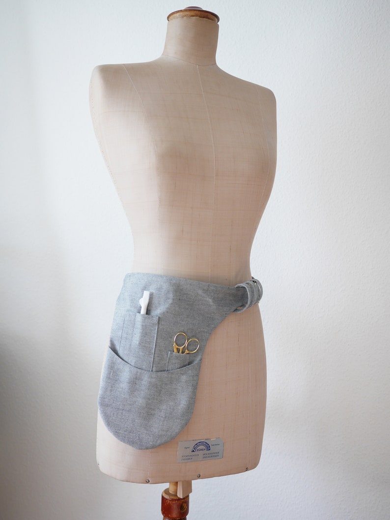 Puede incluir: Un cintur&oacute;n de herramientas de tela gris con un bolsillo y un par de tijeras. El cintur&oacute;n se lleva en un maniqu&iacute;.