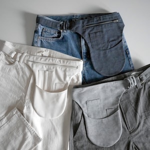 Puede incluir: Cuatro pares de pantalones con una bolsa de tela unida a la cintura. Las bolsas son de diferentes colores: azul oscuro, blanco, gris y negro. Los pantalones son de diferentes colores y telas: mezclilla azul, algod&oacute;n blanco, rayas grises y negro.