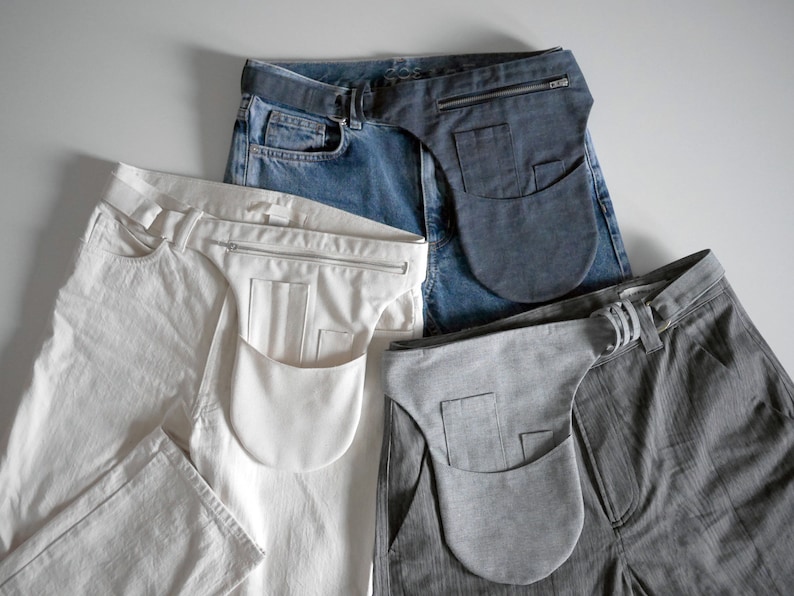 Puede incluir: Cuatro pares de pantalones con una bolsa de tela unida a la cintura. Las bolsas son de diferentes colores: azul oscuro, blanco, gris y negro. Los pantalones son de diferentes colores y telas: mezclilla azul, algod&oacute;n blanco, rayas grises y negro.