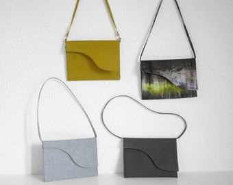 Puede incluir: Cuatro bolsos de hombro de varios colores cuelgan contra una pared blanca. Los bolsos incluyen amarillo mostaza, un estampado negro y verde, azul claro y gris oscuro. Cada bolso tiene un diseño de solapa curvada único y una correa fina.