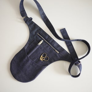 Puede incluir: Un cintur&oacute;n de herramientas de mezclilla azul oscuro con un bolsillo con cremallera y una correa. El cintur&oacute;n tiene un bolsillo con un par de tijeras doradas y un bol&iacute;grafo plateado en su interior.