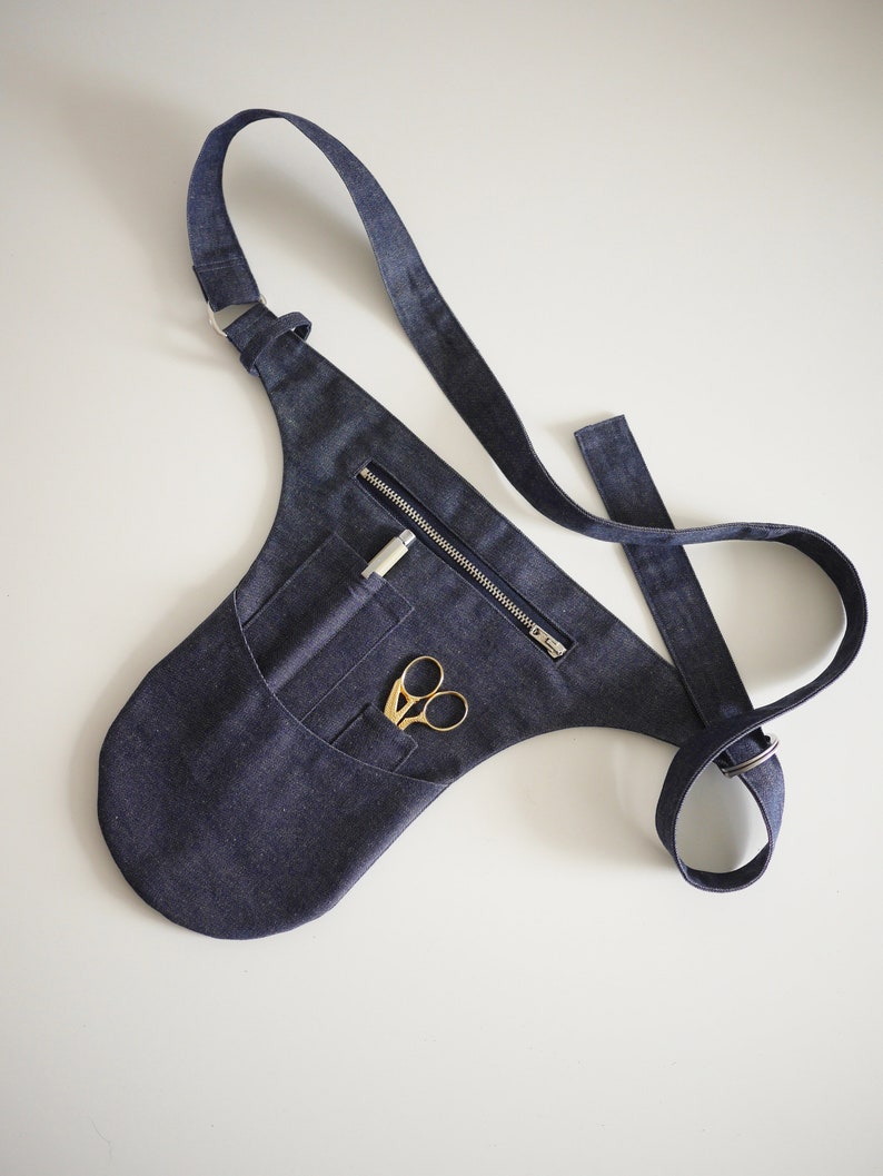 Puede incluir: Un cintur&oacute;n de herramientas de mezclilla azul oscuro con un bolsillo con cremallera y una correa. El cintur&oacute;n tiene un bolsillo con un par de tijeras doradas y un bol&iacute;grafo plateado en su interior.