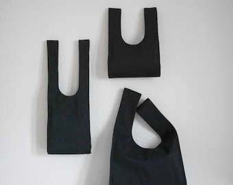 Puede incluir: Tres bolsas de mano negras de diferentes tamaños se muestran contra una pared blanca. Las bolsas tienen un diseño simple y minimalista con una abertura en forma de U y asas integradas. Las bolsas están hechas de una tela oscura y lisa.